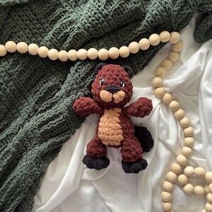May include: A handmade crochet beaver plush toy in shades of brown, tan, and black, with a dark green textured blanket and a wooden bead garland. The beaver has a cute, friendly expression.