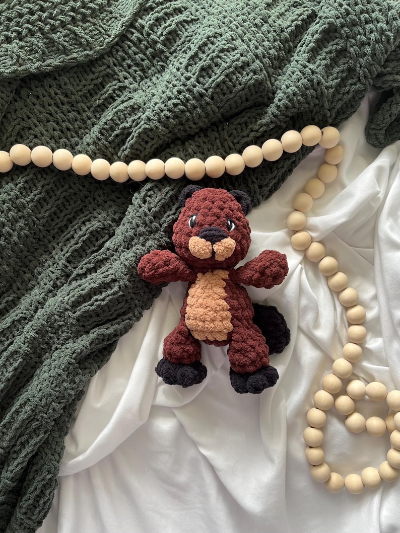 May include: A handmade crochet beaver plush toy in shades of brown, tan, and black, with a dark green textured blanket and a wooden bead garland. The beaver has a cute, friendly expression.