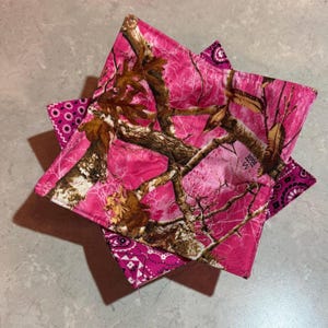 May include: Stack of pink camo fabric bowl cozies. The fabric features a pink camouflage pattern with brown branches and leaves. The edges are trimmed with a paisley print in shades of pink and white. The cozies are square and folded.
