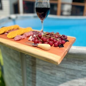 May include: A wooden serving board with a selection of food and a glass of red wine. The board holds crackers, sliced meats, and grapes. A small fork with a wooden handle rests on the board. The background shows a swimming pool.