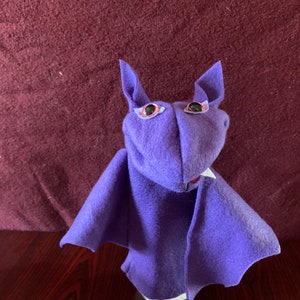 May include: Purple felt hand puppet shaped like a bat with large black eyes and a white mouth.
