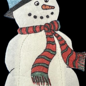 May include: A handmade snowman decoration with a black top hat, blue band, and a red and green striped scarf. The snowman has black dot eyes, a carrot nose, and a stitched outline. A festive winter holiday decoration.