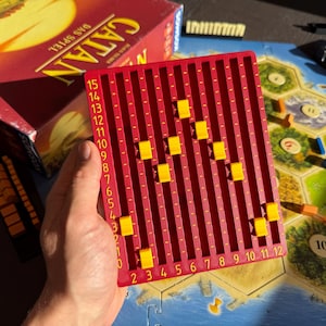 Könnte beinhalten: Ein rotes Kunststoff-Spielbrett zur Punkteverfolgung mit vertikalen Spalten von null bis fünfzehn und horizontalen Reihen von zwei bis zwölf, das kleine gelbe Schieberegler in den vertieften Rillen enthält.