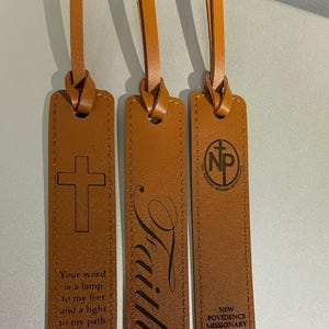 Könnte beinhalten: Drei braune Leder-Lesezeichen mit unterschiedlichen Designs. Das erste Lesezeichen hat ein Kreuz und den Text "Your word is a lamp to my feet and a light to my path Psalm 119:105". Das zweite Lesezeichen hat das Wort "Faith" in Kursivschrift. Das dritte Lesezeichen hat ein Kreuz in einem Kreis mit den Buchstaben "NP" und dem Text "New Providence Missionary Baptist Church".