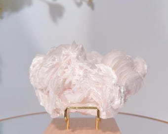 May include: A light pink, textured, crystalline mineral specimen displayed on a small, natural wood block stand with gold-colored metal supports. The background is a soft, blurred white.