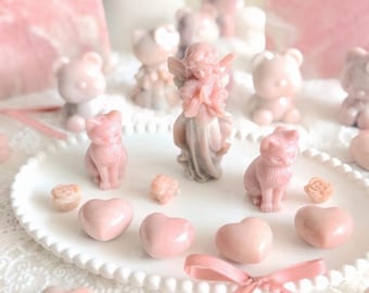 May include: A collection of small, light pink carved stone figurines including an angel, cats, bears, and hearts arranged on a white scalloped tray. The smooth, polished items feature soft pastel tones and detailed shapes, accented by a pink ribbon.