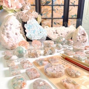 May include: A collection of polished flower agate stones in various shapes including spheres, cabochons, and rectangular slabs, displayed alongside two large raw geode clusters and a calligraphy brush resting on a tan paper with black ink text.