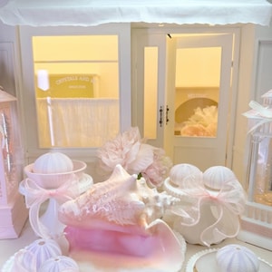 May include: A display of decorative items, including a large pink conch shell, white ceramic pieces, and sea urchins, all adorned with pink ribbons. The shop name "Crystals and Me" is visible above the entrance.