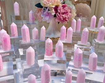 May include: A collection of numerous pink and white crystal points, each shaped into a faceted tower. These stone towers are displayed individually on clear acrylic square pedestals of varying heights, arranged in a group with a floral bouquet.