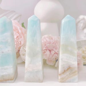 May include: Six light blue and white striped crystal points, also known as celestite, arranged in a row on a white surface. The points are polished and have a smooth, glassy finish.