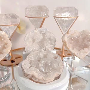 May include: A collection of quartz crystal geodes and points in various shapes, including heart-shaped and diamond-shaped, displayed on decorative stands. The crystals are a pale, translucent color, and the stands are gold and silver.