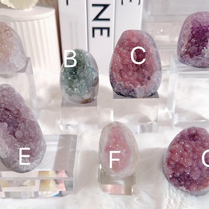Puede incluir: Siete geodas de amatista diferentes, cada una con un color y un patrón únicos. Las geodas se muestran en pequeños soportes transparentes. Las geodas están etiquetadas con las letras A a G.