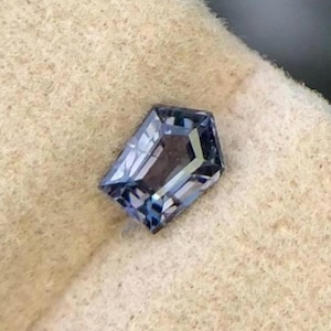 Könnte beinhalten: Ein facettierter, blauer Edelstein mit rechteckiger Form und abgeschrägten Ecken. Der Stein hat eine reflektierende Oberfläche, die innere Facetten und eine Reihe von Blautönen zeigt. Der Edelstein ist vor einem neutralen, strukturierten Hintergrund platziert.