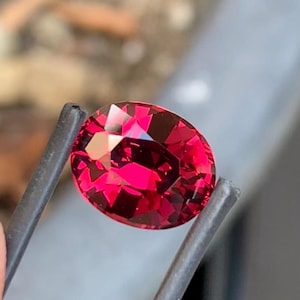 Puede incluir: Una gema de corte ovalado vibrante con un color rojo profundo y saturado y una superficie multifacetada que refleja la luz. La piedra se sostiene de forma segura entre las dos puntas metálicas oscuras y delgadas de unas pinzas.