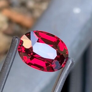Puede incluir: Una gema roja de corte ovalado con una superficie facetada que refleja la luz, creando reflejos carmesí profundos y escarlatas brillantes. La piedra está sujeta por dos finas pinzas metálicas oscuras, mostrando su claridad y color intenso.