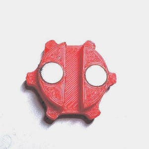 May include: A red plastic 3D printed object with two white circles. The object has a textured surface and a geometric shape.