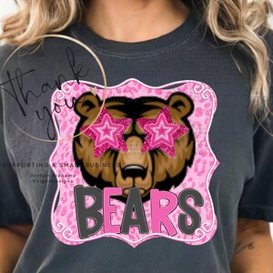 Puede incluir: Camiseta gris oscuro con un oso pardo que lleva gafas de sol rosas en forma de estrella. El gráfico del oso está sobre un fondo rosa con estampado de leopardo y la palabra "BEARS" en rosa y gris.