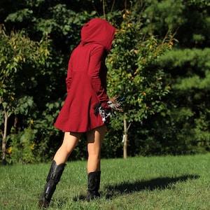 May include: A woman wearing a red hooded tunic with a long, flowing skirt. She is standing in a grassy field with trees in the background.