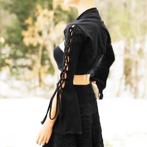 May include: Black cropped top with long sleeves featuring lace-up detailing on the arms. The top has a fitted bodice and a slightly flared hem.