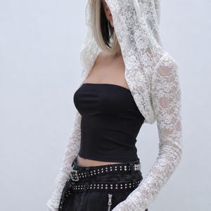 May include: A white lace hooded shrug with long sleeves. The shrug is paired with a black tube top and a studded belt. The outfit is set against a plain white background.