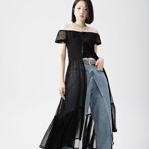 May include: A woman wearing a black sheer off-the-shoulder dress with a ruffled hem over a pair of blue denim jeans. She is wearing a silver chain necklace and a black belt with a silver buckle.