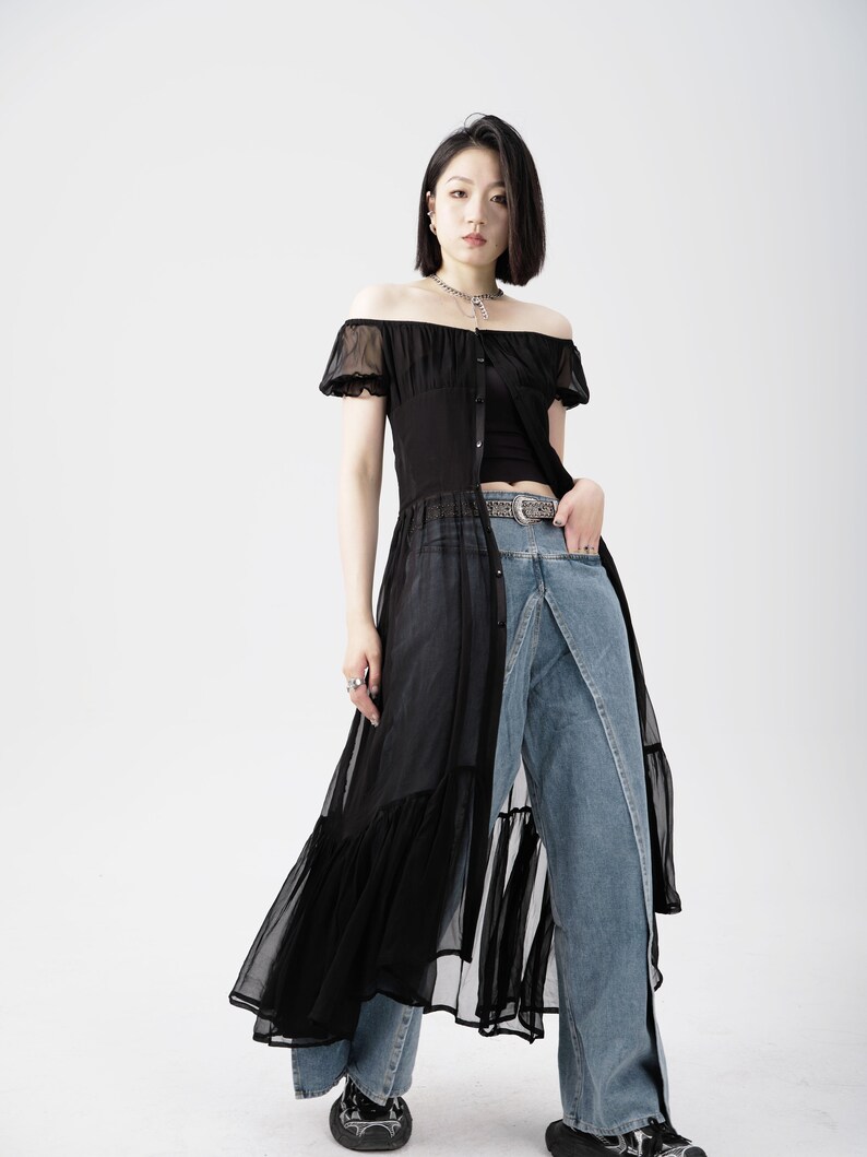 May include: A woman wearing a black sheer off-the-shoulder dress with a ruffled hem over a pair of blue denim jeans. She is wearing a silver chain necklace and a black belt with a silver buckle.