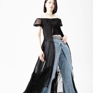 May include: A woman wearing a black sheer off-the-shoulder dress with a ruffled hem and a pair of blue denim jeans. She is wearing a silver chain necklace and a silver belt.