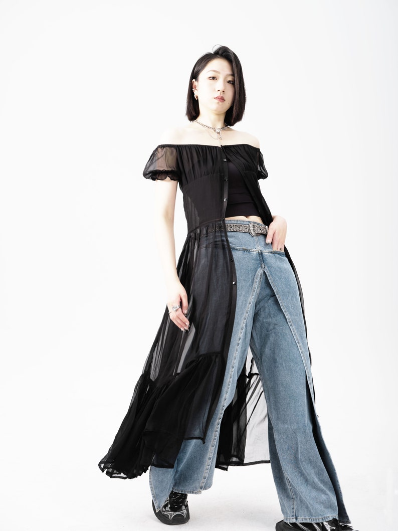 May include: A woman wearing a black sheer off-the-shoulder dress with a ruffled hem and a pair of blue denim jeans. She is wearing a silver chain necklace and a silver belt.