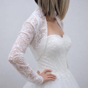 May include: A white lace bolero jacket with long sleeves and a floral pattern, worn over a strapless white gown featuring a sweetheart neckline and intricate lace embroidery on the bodice. The bolero has a cropped cut and scalloped edges.