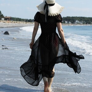 May include: A woman wearing a black sheer dress with a white straw hat walks along a beach.