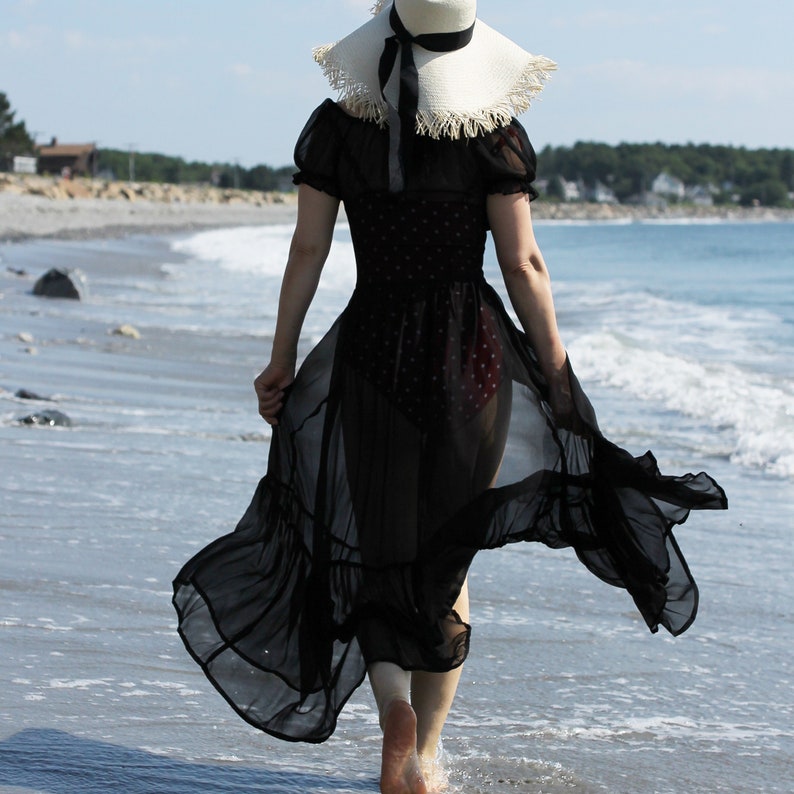 May include: A woman wearing a black sheer dress with a white straw hat walks along a beach.