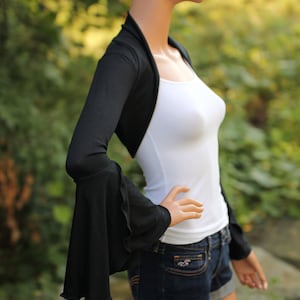 Könnte beinhalten: Schwarze Bolerojacke mit langen Glockenärmeln. Die Bolerojacke ist aus einem weichen, dehnbaren Stoff gefertigt und hat einen drapierten Ausschnitt.