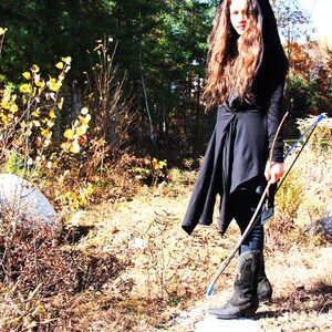 May include: A woman wearing a black hooded cloak and long brown hair holds a wooden bow and arrow in a forest setting. The background features fall foliage in shades of yellow, orange, and red.