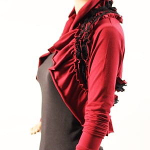 May include: A burgundy bolero jacket with long sleeves and a draped, ruffled design. Black lace trim accents the neckline and front. The jacket is worn over a black top, creating a layered look. The fabric appears soft and flowing.