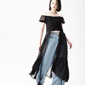 May include: A woman wearing a black sheer off-the-shoulder dress over a black tank top and blue jeans. She is wearing a silver chain necklace and a silver belt.