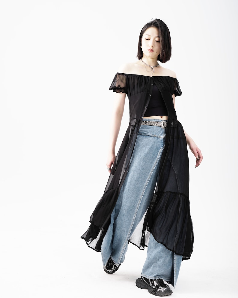May include: A woman wearing a black sheer off-the-shoulder dress over a black tank top and blue jeans. She is wearing a silver chain necklace and a silver belt.