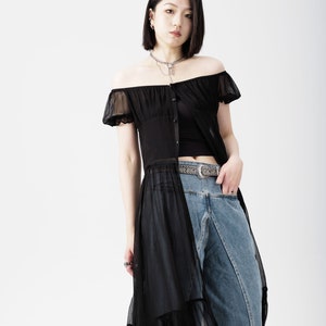 May include: A woman wearing a black off-the-shoulder sheer top with a black tank top underneath. She is wearing blue jeans with a silver belt and has a silver chain necklace.