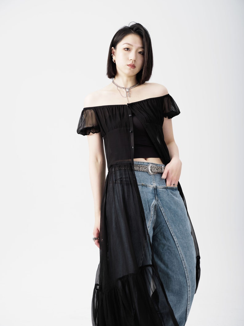 May include: A woman wearing a black off-the-shoulder sheer top with a black tank top underneath. She is wearing blue jeans with a silver belt and has a silver chain necklace.