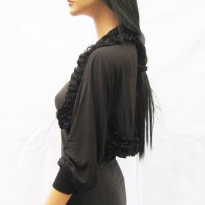 Könnte beinhalten: Schwarze Bolerojacke mit Rüschenbesatz. Die Bolerojacke hat lange Ärmel und ist aus einem weichen, dehnbaren Stoff gefertigt.