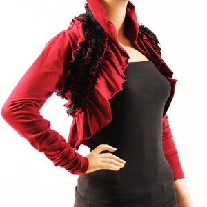 May include: A burgundy bolero with long sleeves and a ruffled neckline. The bolero has black lace trim around the neckline and shoulders.