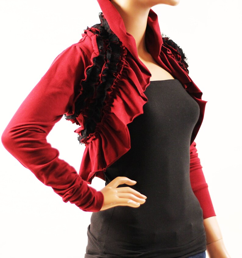 May include: A burgundy bolero with long sleeves and a ruffled neckline. The bolero has black lace trim around the neckline and shoulders.