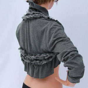 May include: A gray cropped cardigan with a ruffled lace trim. The cardigan has long sleeves and a relaxed fit.