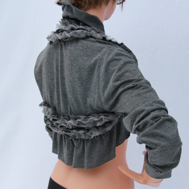 May include: A gray cropped cardigan with a ruffled lace trim. The cardigan has long sleeves and a relaxed fit.