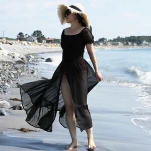 May include: A woman wearing a black sheer maxi dress with a ruffled hem and a white wide-brimmed hat. She is standing on a sandy beach with the ocean in the background.