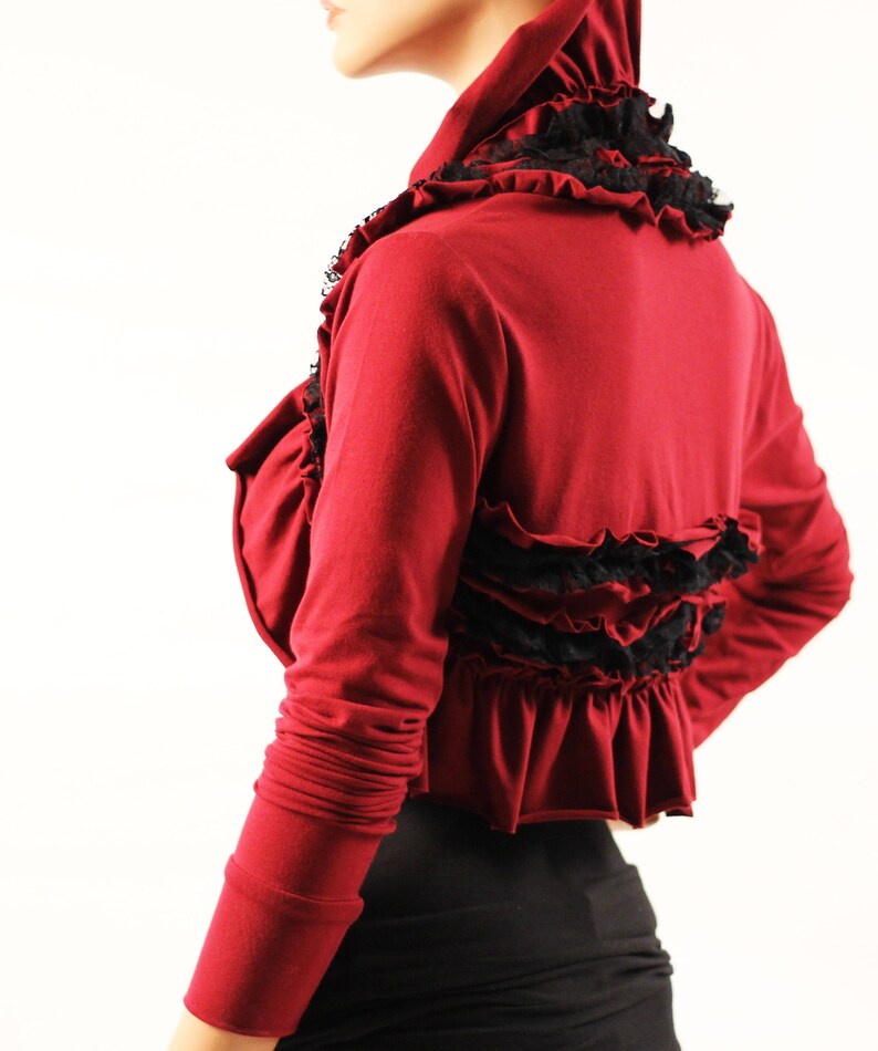 May include: A maroon long-sleeved shrug with black lace trim and ruffles. The shrug has a high neckline and a cropped length.