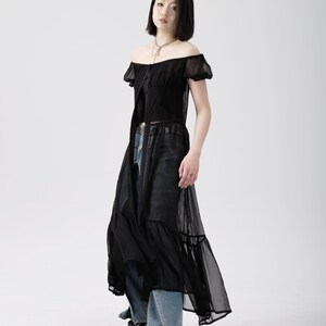 May include: A black sheer maxi dress with a ruffled hem and short puff sleeves. The dress has a button-up front and a fitted bodice. The model is wearing the dress with blue jeans and black platform shoes.