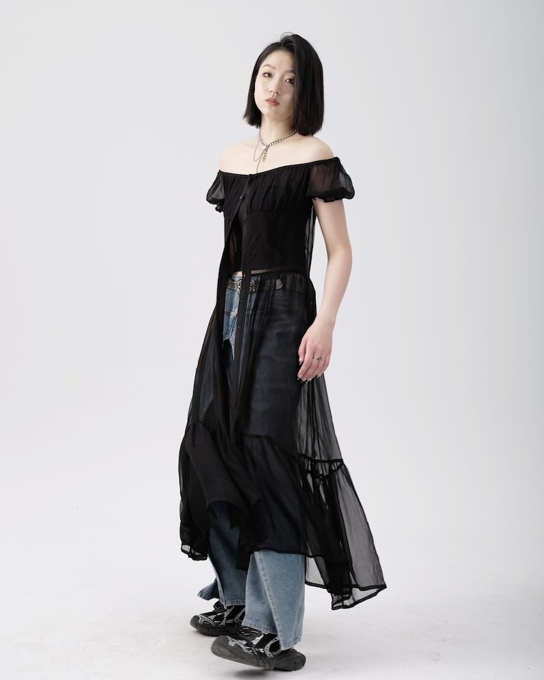 May include: A black sheer maxi dress with a ruffled hem and short puff sleeves. The dress has a button-up front and a fitted bodice. The model is wearing the dress with blue jeans and black platform shoes.
