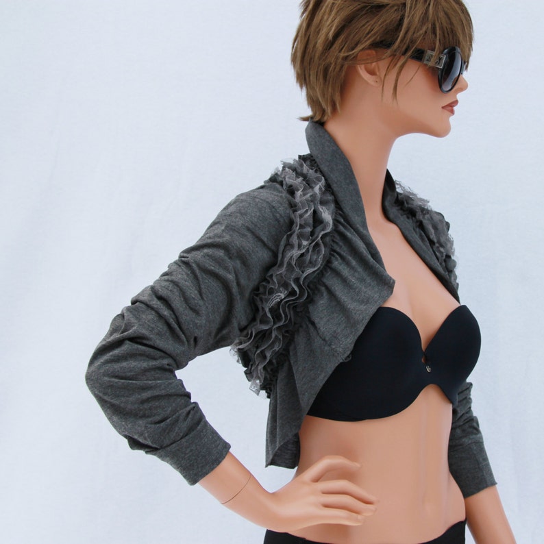 May include: A gray cropped cardigan with a ruffled trim and a black bra underneath. The cardigan has long sleeves and a draped neckline.