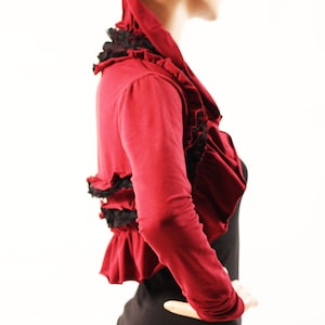 May include: A red cropped cardigan with black lace trim and ruffles. The cardigan has long sleeves and a scoop neck.