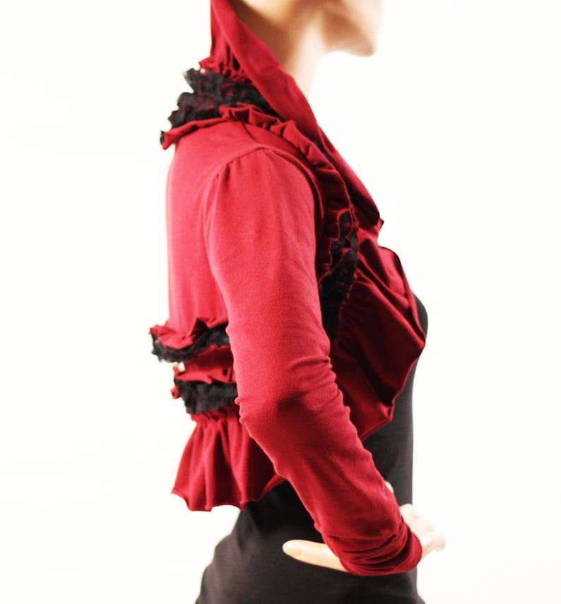 May include: A red cropped cardigan with black lace trim and ruffles. The cardigan has long sleeves and a scoop neck.